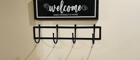 Un letrero montado en la pared con la inscripción 'welcome make yourself at home' sobre un perchero negro fijado a la pared con ganchos. Abajo, un cojín blanco con textura descansa sobre una superficie de madera.