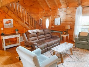 A cozy cabin interior with a brown leather sofa, a white armchair, a wooden coffee table, and a staircase leading to an upper level.