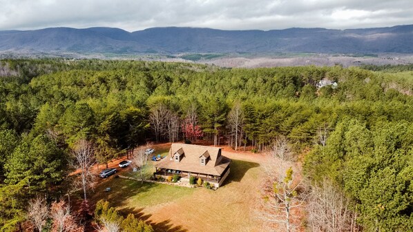 A large, single-story house surrounded by dense forest, with a clear view of distant mountains.