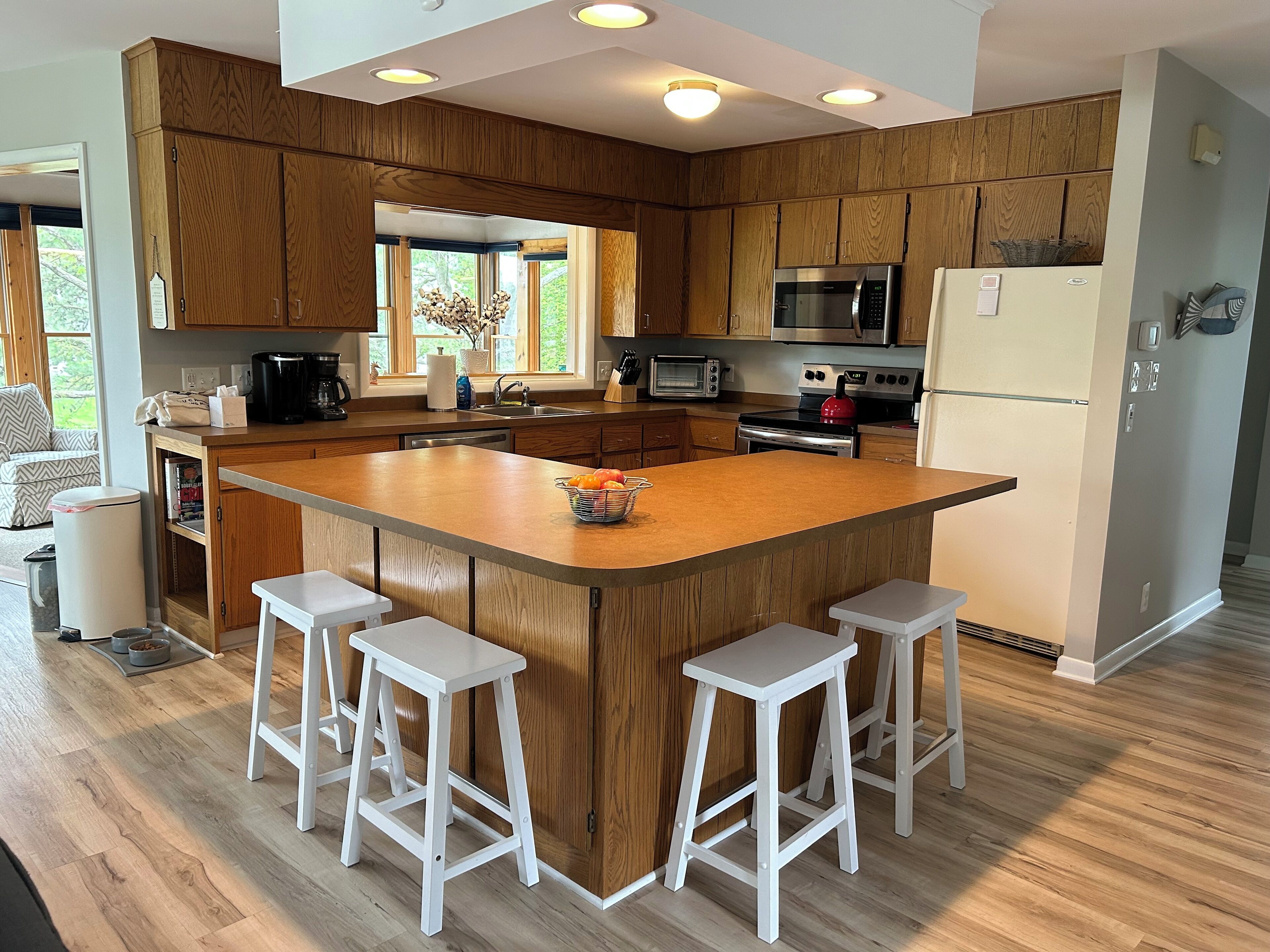 Una cocina moderna con armarios de madera, una isla central con taburetes, electrodomésticos de acero inoxidable y un amplio ventanal que proporciona luz natural.