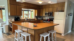 Una cocina moderna con armarios de madera, una isla central con taburetes, electrodomésticos de acero inoxidable y un amplio ventanal que proporciona luz natural.