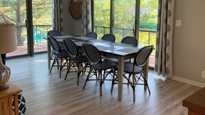 Un comedor con una mesa grande y seis sillas, ventanas amplias con cortinas, y una lámpara colgante.