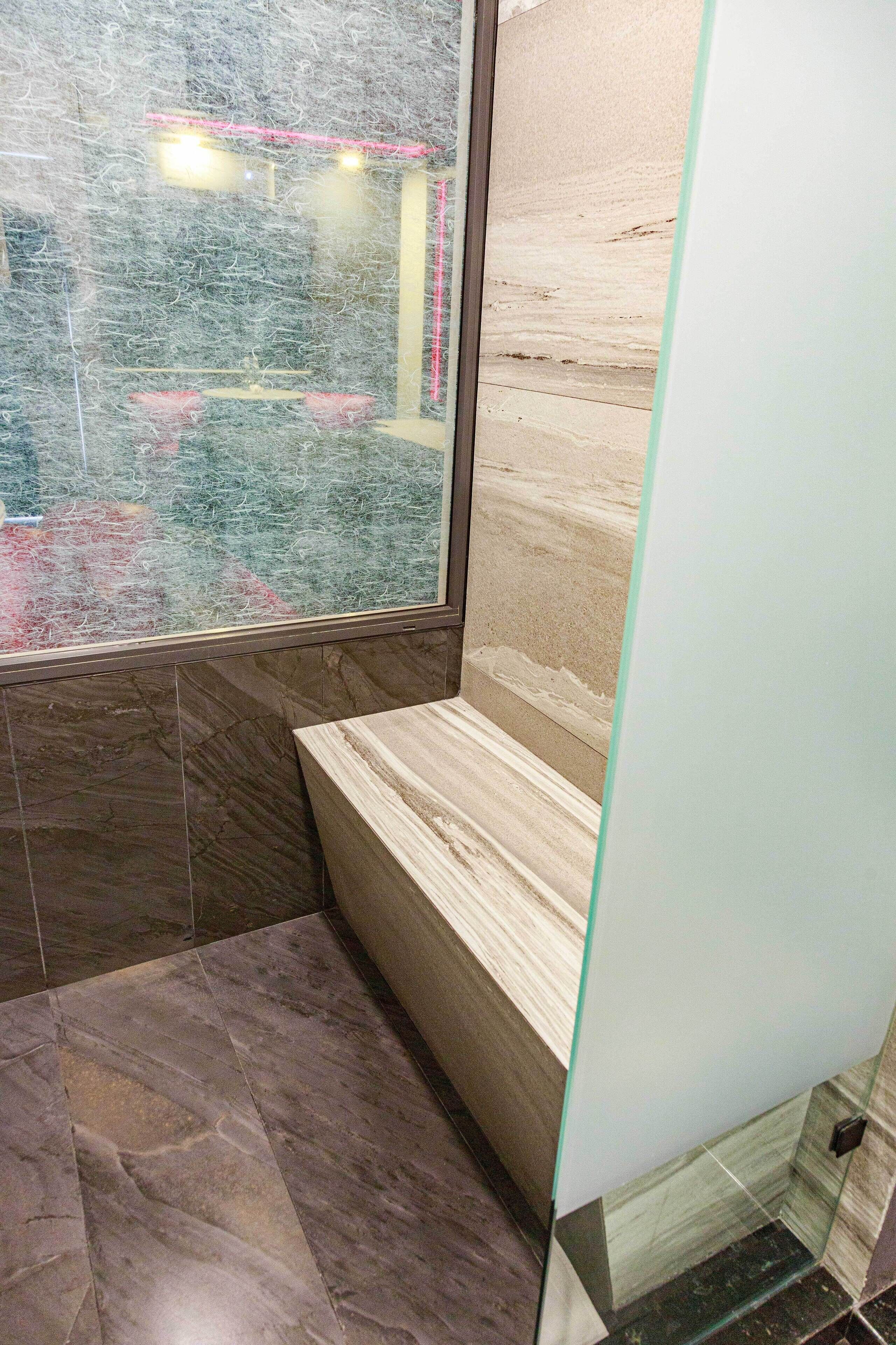 Un baño con cabina de ducha de vidrio, azulejos de mármol y un banco.
