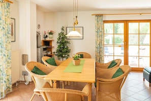 Een eetgedeelte met een houten tafel en rieten stoelen, een potplant en uitzicht op het omliggende landschap door een glazen deur.