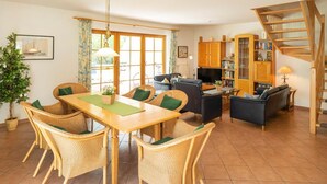 A spacious living area with a dining table, wicker chairs, a sofa, and a bookshelf. - Spacious semi-detached house with garden, terrace and pool in the Zingst vacation park - (Zingst)