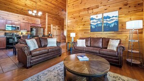 L'intérieur d'un chalet en bois comprend deux canapés en cuir, une table basse ronde, un coin cuisine et un tableau accroché au mur.