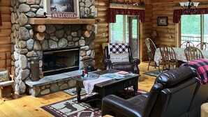 A cozy cabin interior with a stone fireplace, wooden walls, a ceiling fan, and a dining area.