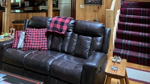A cabin interior with a wooden staircase, a dining area, a kitchen, and a living room with a leather couch and plaid cushions.