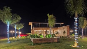 Un edificio moderno con fachada de vidrio, rodeado por un jardín iluminado con palmeras y un área pavimentada.
