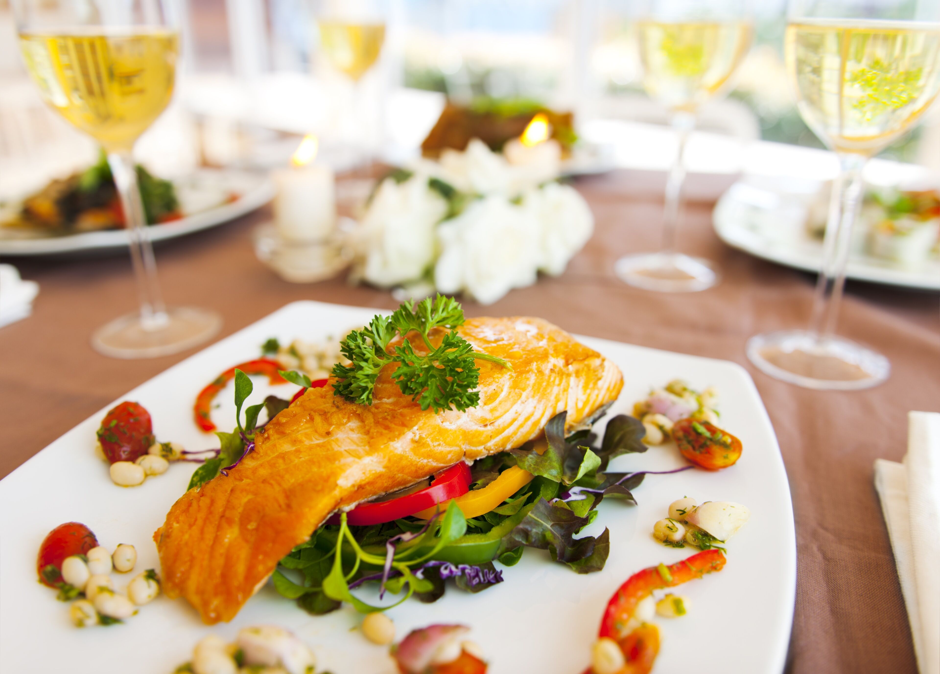 A salmon dish on a white plate with greens and vegetables, surrounded by wine glasses and candles.