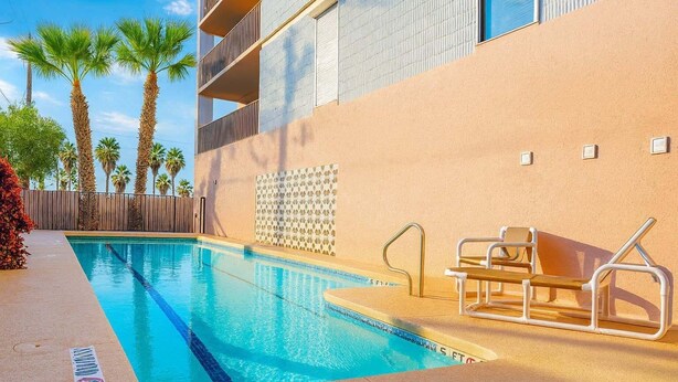 A swimming pool with a lifeguard chair and a pool ladder, surrounded by palm trees and a building with a beige facade. - Walk to Beach - Heart of Entertainment District 1 Bedroom Home by RedAwning (South Padre Island)