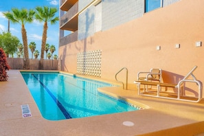 Una piscina con una silla de salvavidas y una escalera, rodeada de palmeras y un edificio con fachada color beige.
