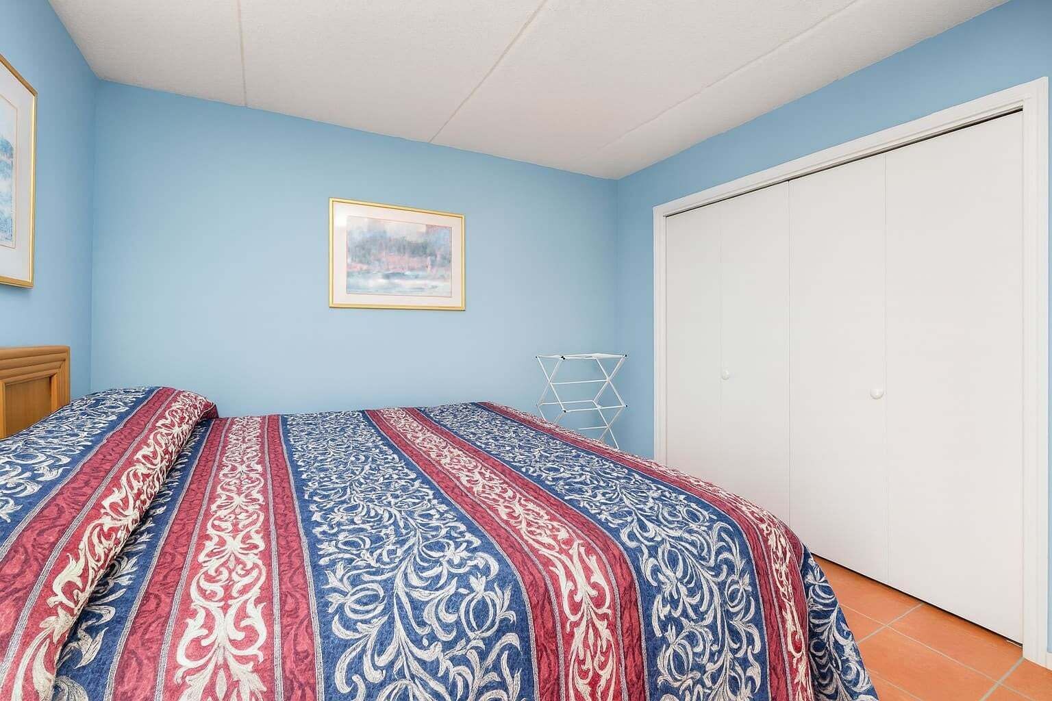 A bedroom with a bed, a white wardrobe, a framed picture on the wall, and a drying rack.