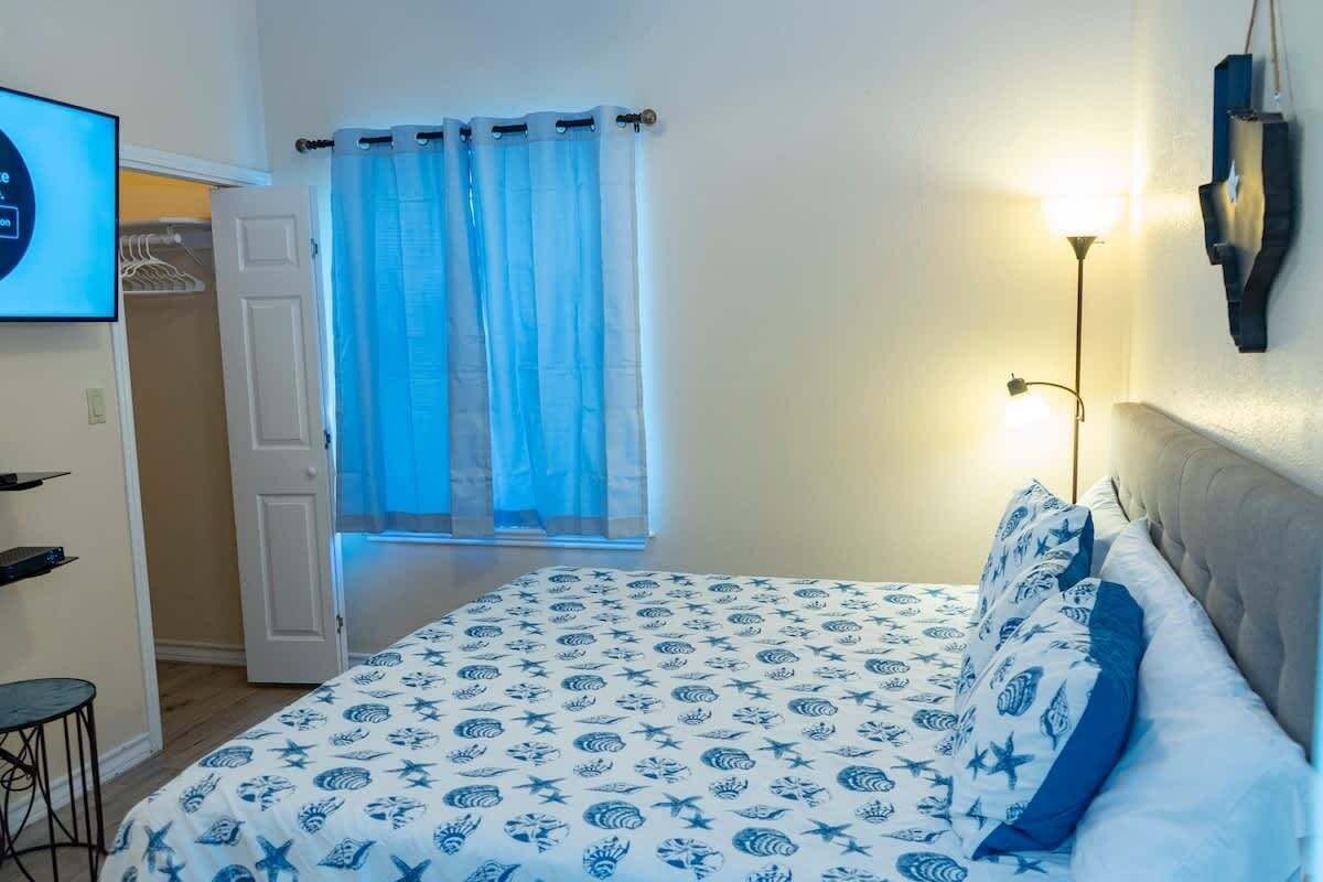 A bedroom with a bed, a bedside table, a chair, a curtain, and a wall-mounted light fixture.