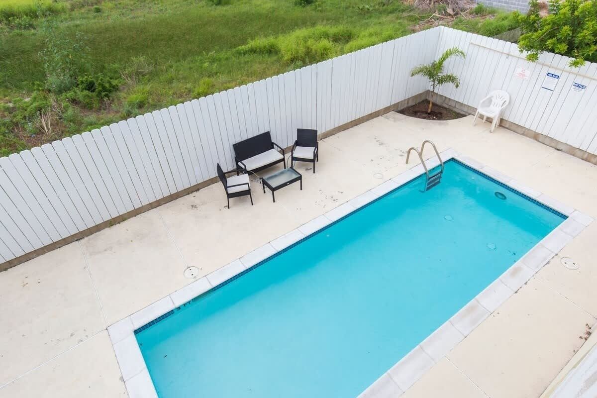 A rectangular swimming pool with a ladder, surrounded by a white fence. There are three chairs and a small table near the pool.