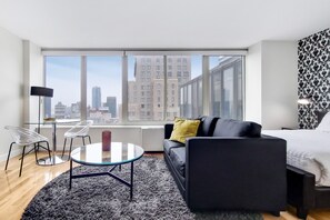 A modern hotel room with a large window, a black sofa, a glass coffee table, and a city view.