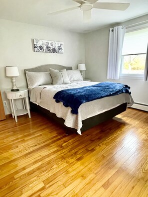 A bedroom with a bed, two bedside tables, a ceiling fan, a window with curtains, and a wall-mounted artwork.