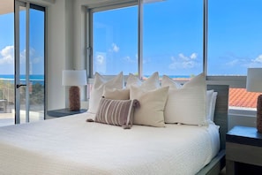 Un dormitorio con una cama grande, varias almohadas, una mesita de noche con lámpara y vistas al océano a través de amplios ventanales.