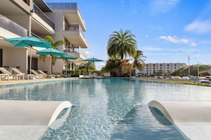 Una piscina con sillas de descanso y sombrillas, un edificio de varios pisos con balcones y palmeras.