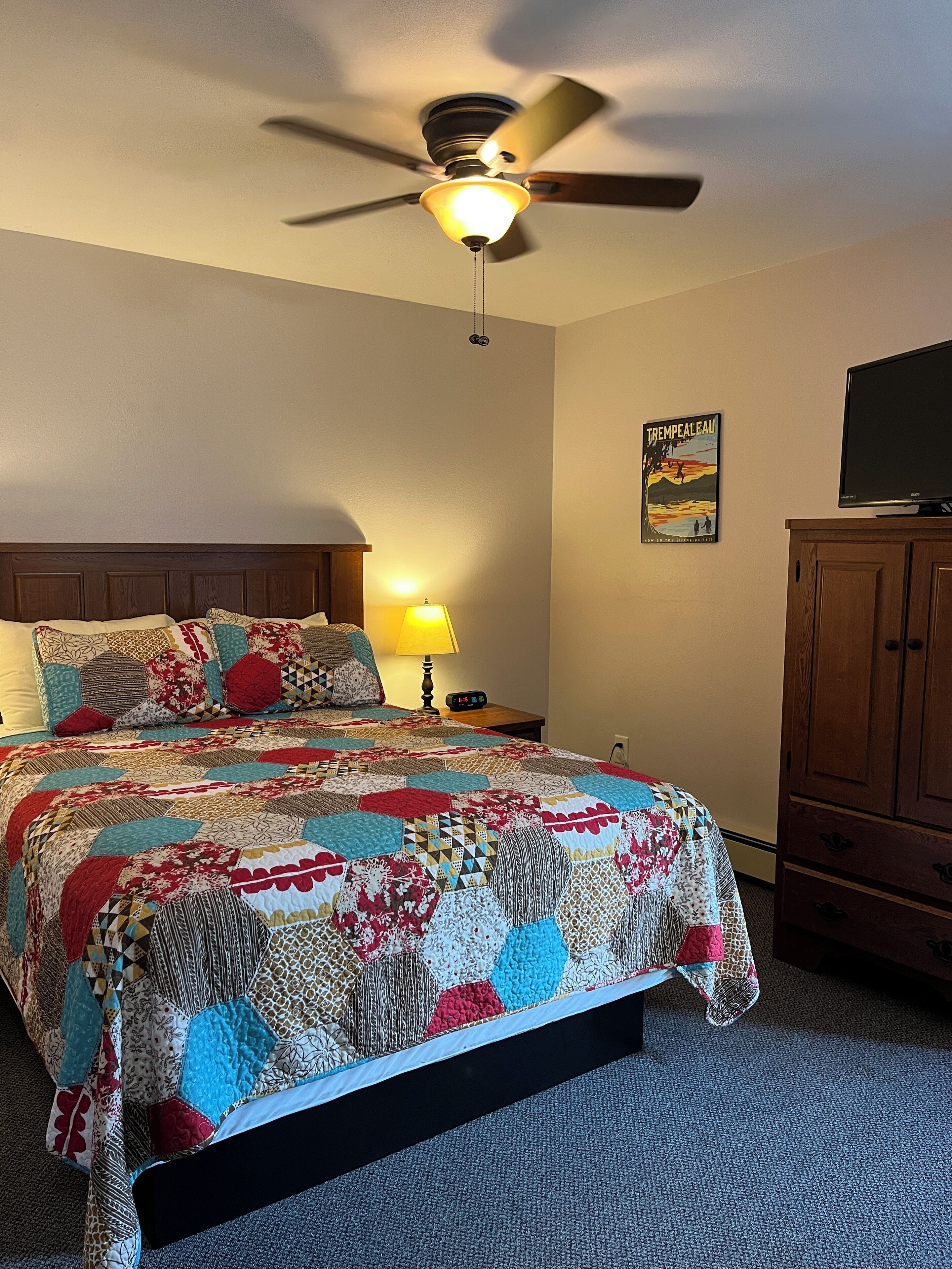 A bedroom with a bed, a ceiling fan, a nightstand with a lamp, a TV on a wooden cabinet, and a poster on the wall.