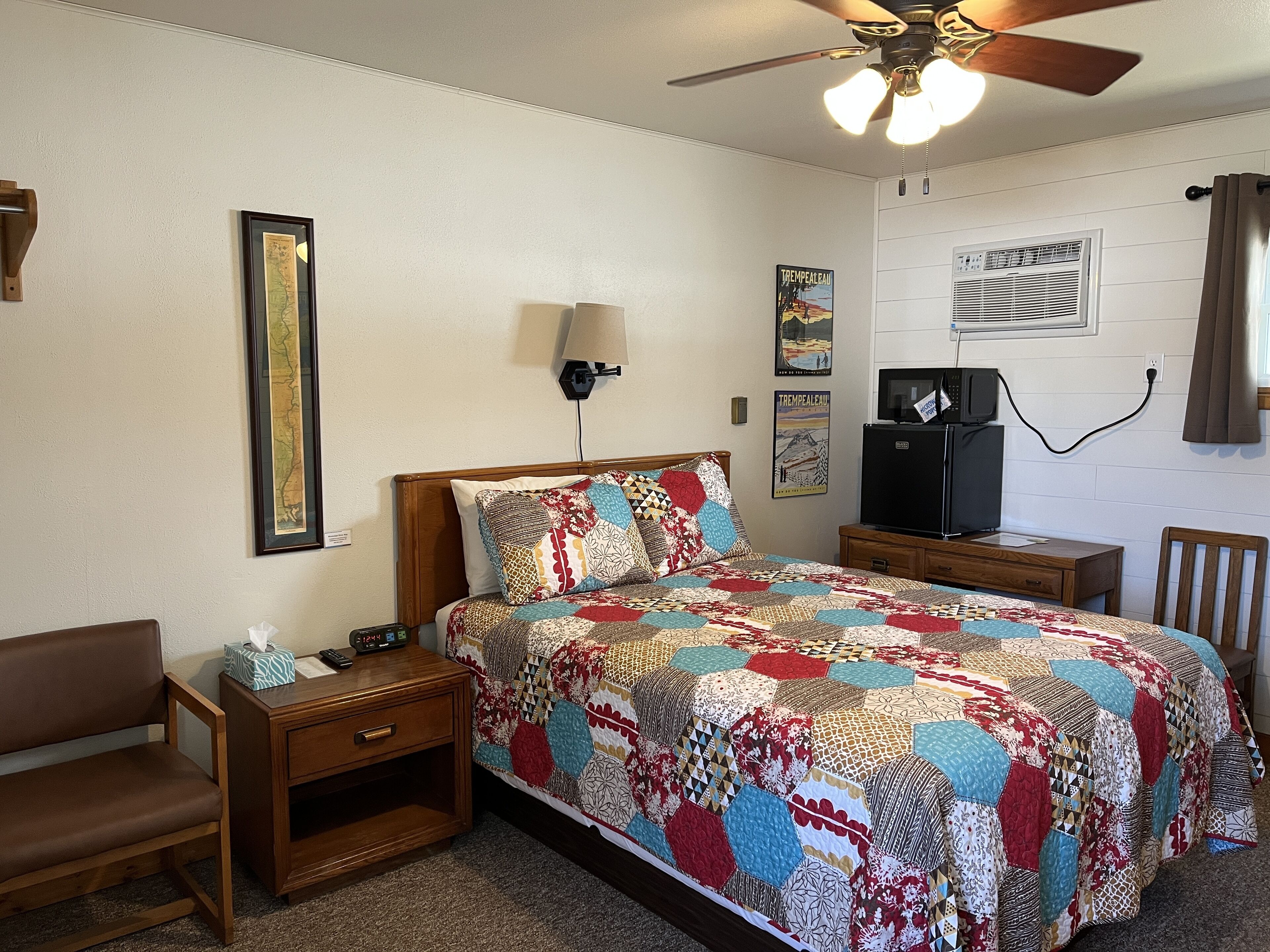 A bedroom with a wooden bed, a nightstand, a desk, a chair, a ceiling fan, and a wall-mounted TV.