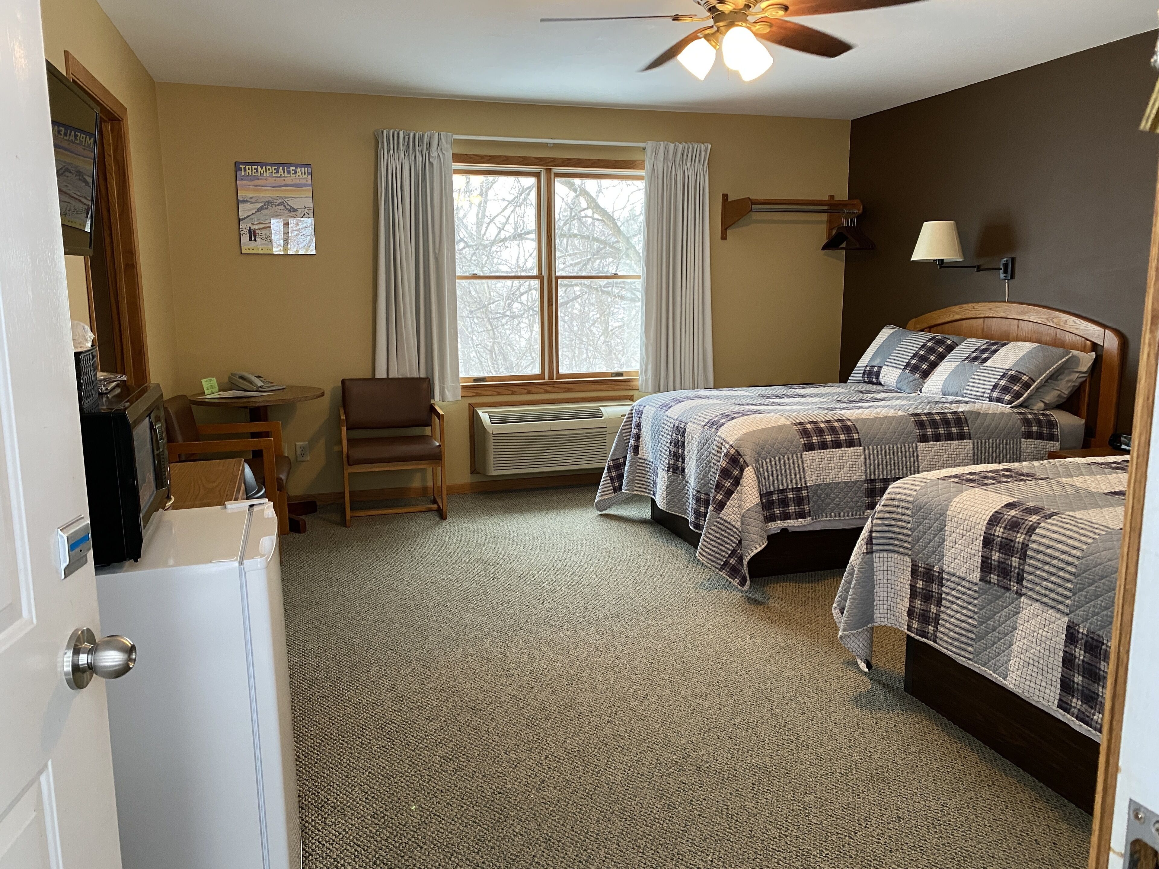 A bedroom with two beds, a desk, a chair, a window with curtains, a ceiling fan, and a wall-mounted lamp.