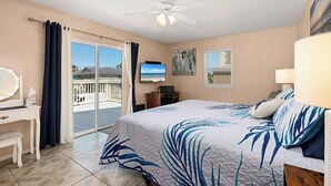 A bedroom with a bed, a desk, a chair, a ceiling fan, and a view of the outside.