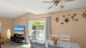 A room with a dining table, chairs, a ceiling fan, a TV, and a view of the beach through a glass door.