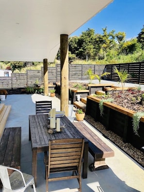 An outdoor patio area with wooden tables and chairs, a pergola, and raised garden beds with plants. - Cablecrest - 2 Master Suites and Grand Views (Cable Bay)