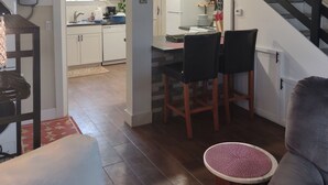 Una cocina moderna con armarios blancos, una zona de comedor con dos sillas, una escalera y una sala de estar con un sofá y una mesa redonda.
