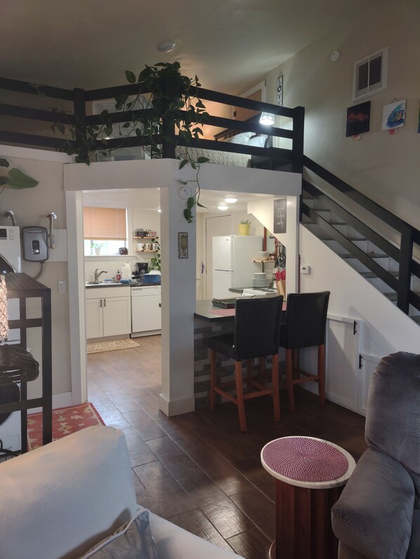 A modern kitchen with white cabinets, a dining area with two chairs, a staircase, and a living room with a sofa and a round table.