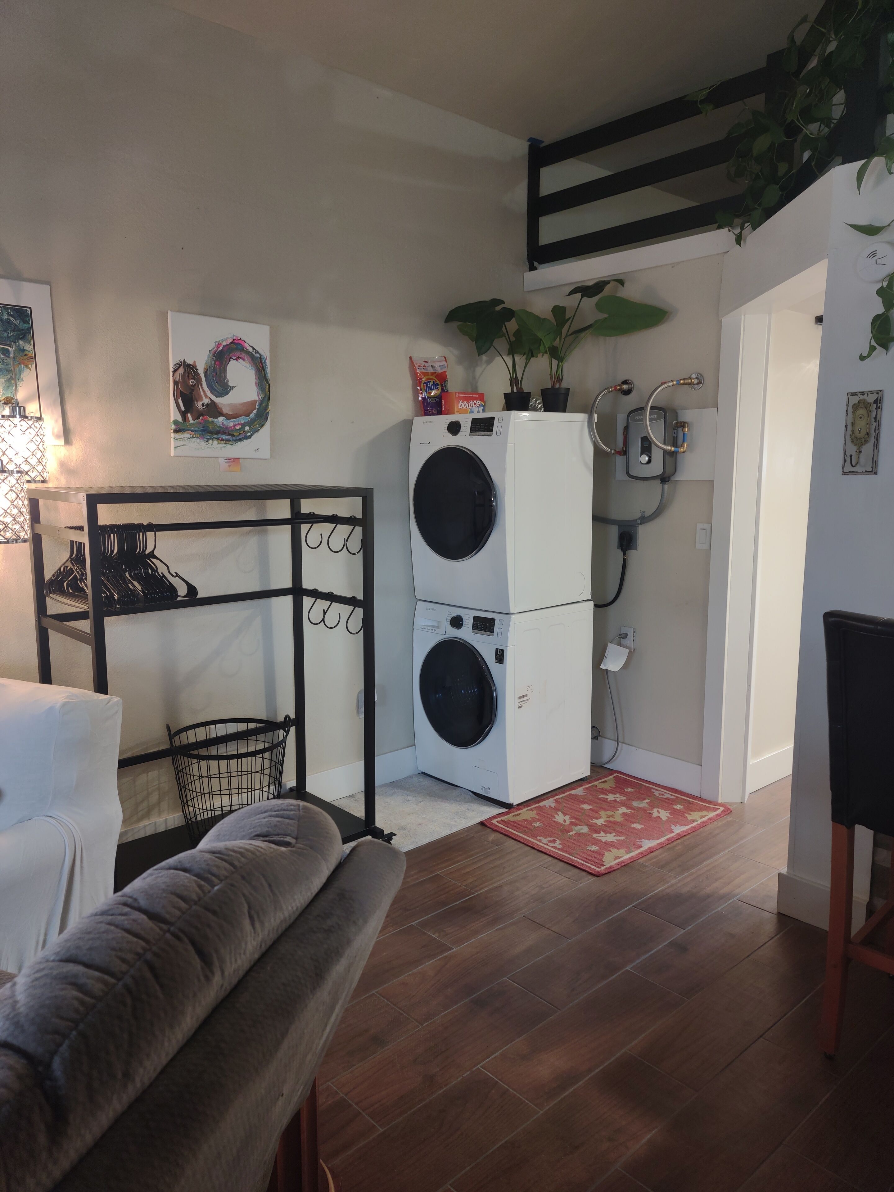 A compact laundry room with a front-loading washer and dryer, a black metal drying rack, a grey couch, and a red rug.