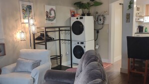 Una sala de estar con un sofá gris, un sillón blanco, una mesa de centro negra y una alfombra. Se ve una lavadora y una planta al fondo.