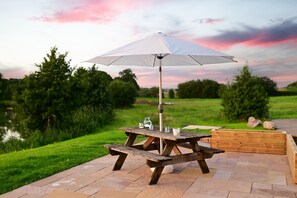 Une table de pique-nique avec un parasol blanc, entourée d’une terrasse en pierre, d’un banc en bois et d’un petit étang.