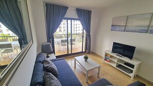 A modern living room with a sofa, a coffee table, a TV on a stand, and a balcony with a view of buildings. - Lovely 2-Bed Apartment in Marina de la Duquesa (Puerto de la Duquesa)