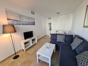 Ein Wohnzimmer mit Fernseher auf einem weißen Schrank, einem dunkelblauen Sofa mit gemusterten Kissen, einem weißen Couchtisch und einer Stehlampe.