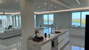Une cuisine moderne dotée d’un grand îlot central, d’un coin repas avec un canapé et de grandes fenêtres offrant une vue sur la ville et l’eau.
