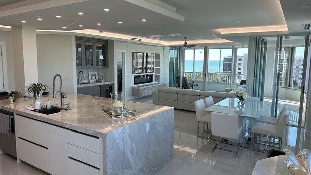 Une cuisine moderne avec un îlot en marbre, un coin repas doté d’une table en verre et un salon avec un canapé et un balcon offrant une vue sur la mer.