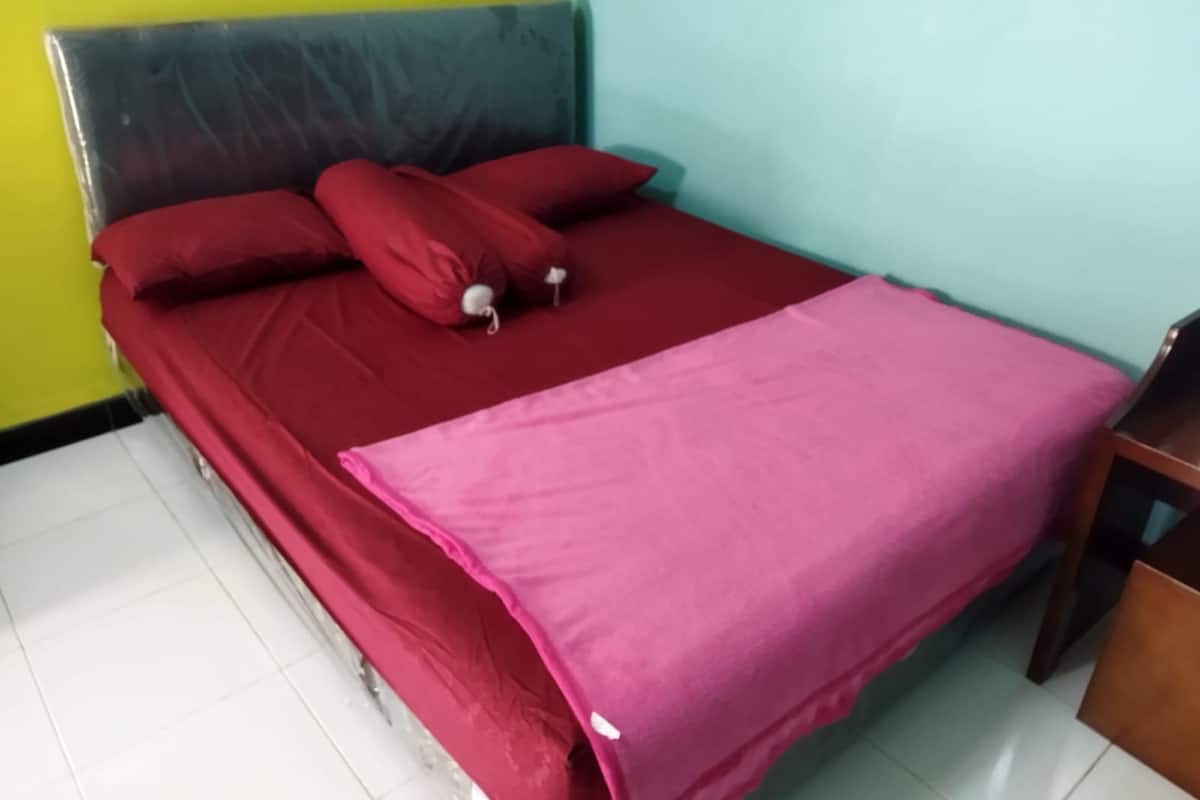 A bed with a red bedspread and pillows, a wooden headboard, and a pink blanket.