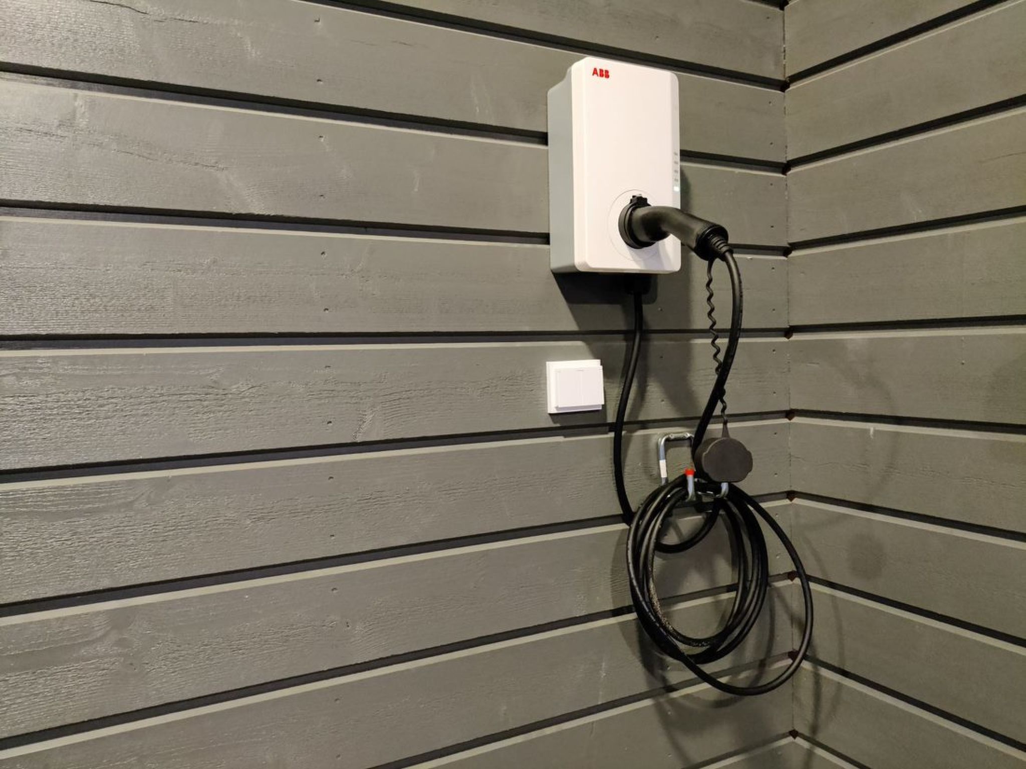 A wall-mounted electrical outlet with a power cord plugged in, set against a grey wall with horizontal paneling.