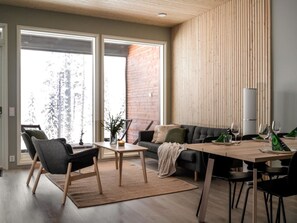 Un salon moderne comprenant un canapé, des fauteuils, une table à manger pour quatre personnes et une grande fenêtre donnant sur un paysage arboré.