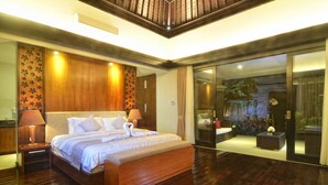 A spacious bedroom with a large bed, wooden headboard, and a sliding glass door leading to a balcony with plants. - Room-kori Maharani Villas - One-bedroom (Gianyar)