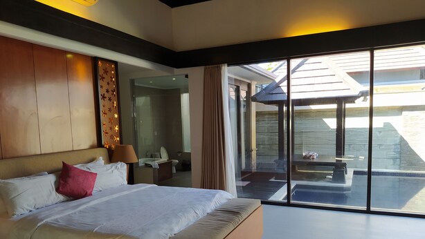 A modern hotel room with a large bed, a bathroom visible through a glass door, and a balcony with a table and chairs. - Room-kori Maharani Villas - One-bedroom (Gianyar)