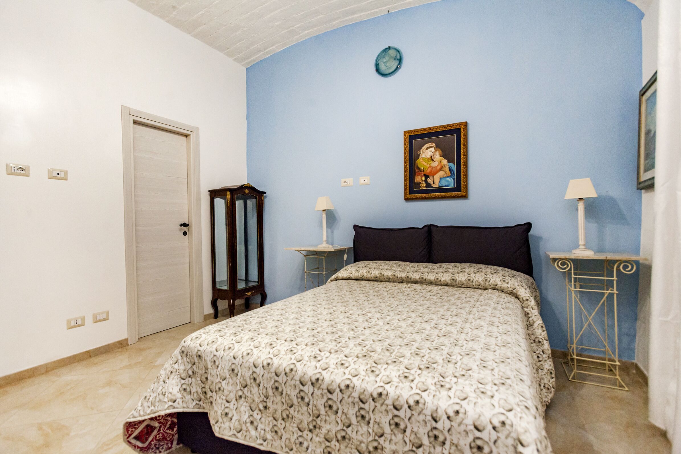 Un dormitorio con una cama, una puerta, un reloj de pared, un cuadro enmarcado y dos mesitas de noche con lámparas.