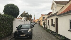 Una estrecha calle empedrada flanqueada por edificios tradicionales, con la aguja de una iglesia a lo lejos y coches aparcados.