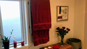 Un baño con una toalla roja colgada en un perchero, un cuadro en la pared, un jarrón con rosas rojas sobre una mesa de madera, y un cubo de basura.