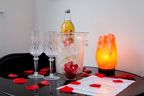 Ein Tisch mit zwei Kristallgläsern, einer Shloer-Flasche, einem Gefäß mit Eis und roten Bonbons, einer Himmalsalzlampe und einer Speisekarte, verziert mit roten Rosenblättern.