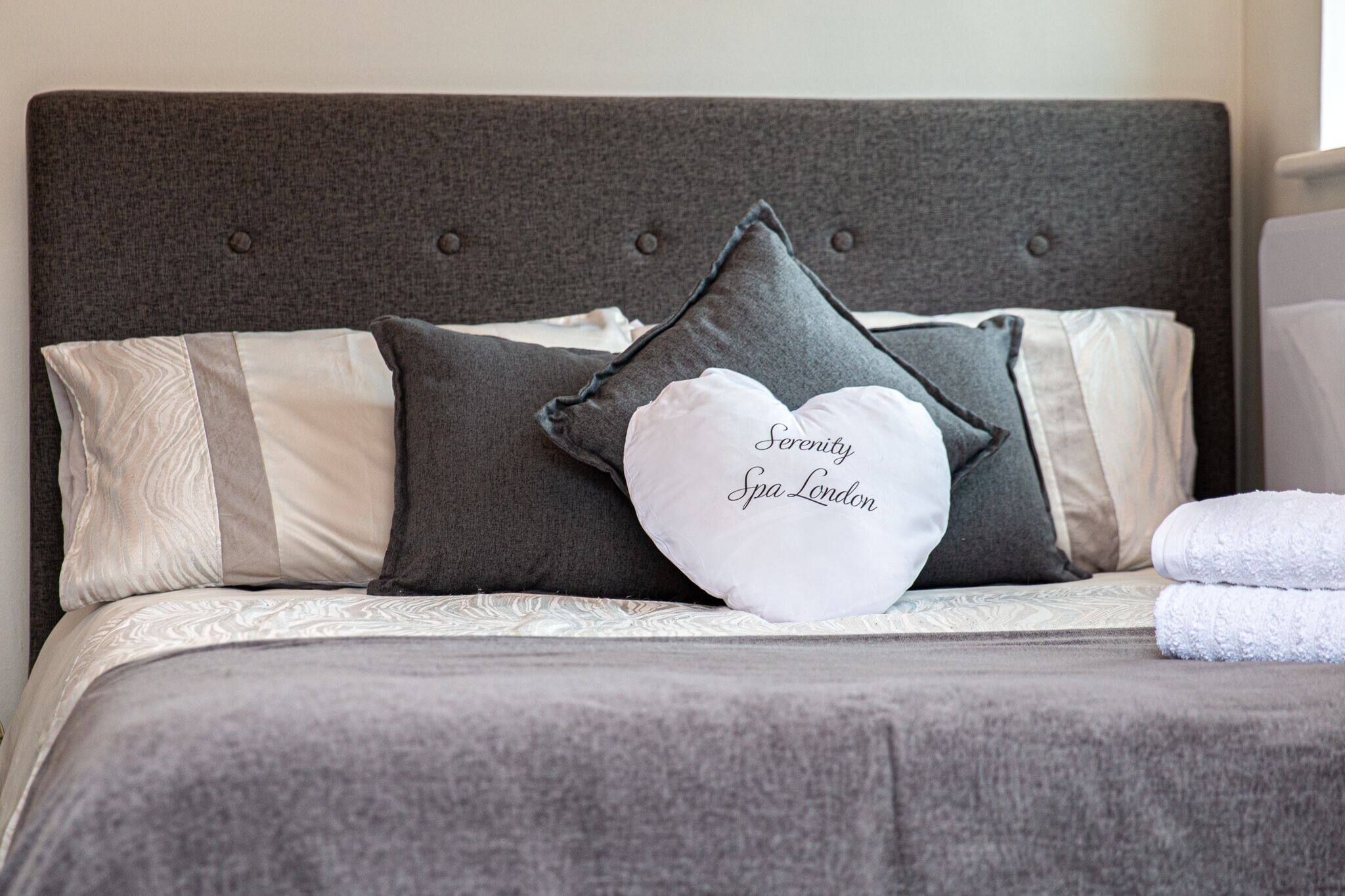 A neatly made bed with a grey headboard, several pillows, and a folded white towel.