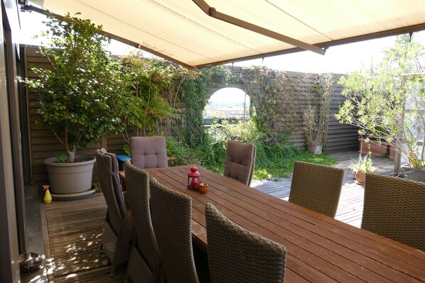 Une terrasse extérieure avec une table en bois et des chaises en osier, entourée de plantes en pot et d’un mur en briques.