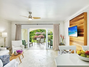 Uma sala de estar moderna com ventilador de teto, TV fixada em uma parede de madeira e vista para um jardim tropical através de portas de vidro.